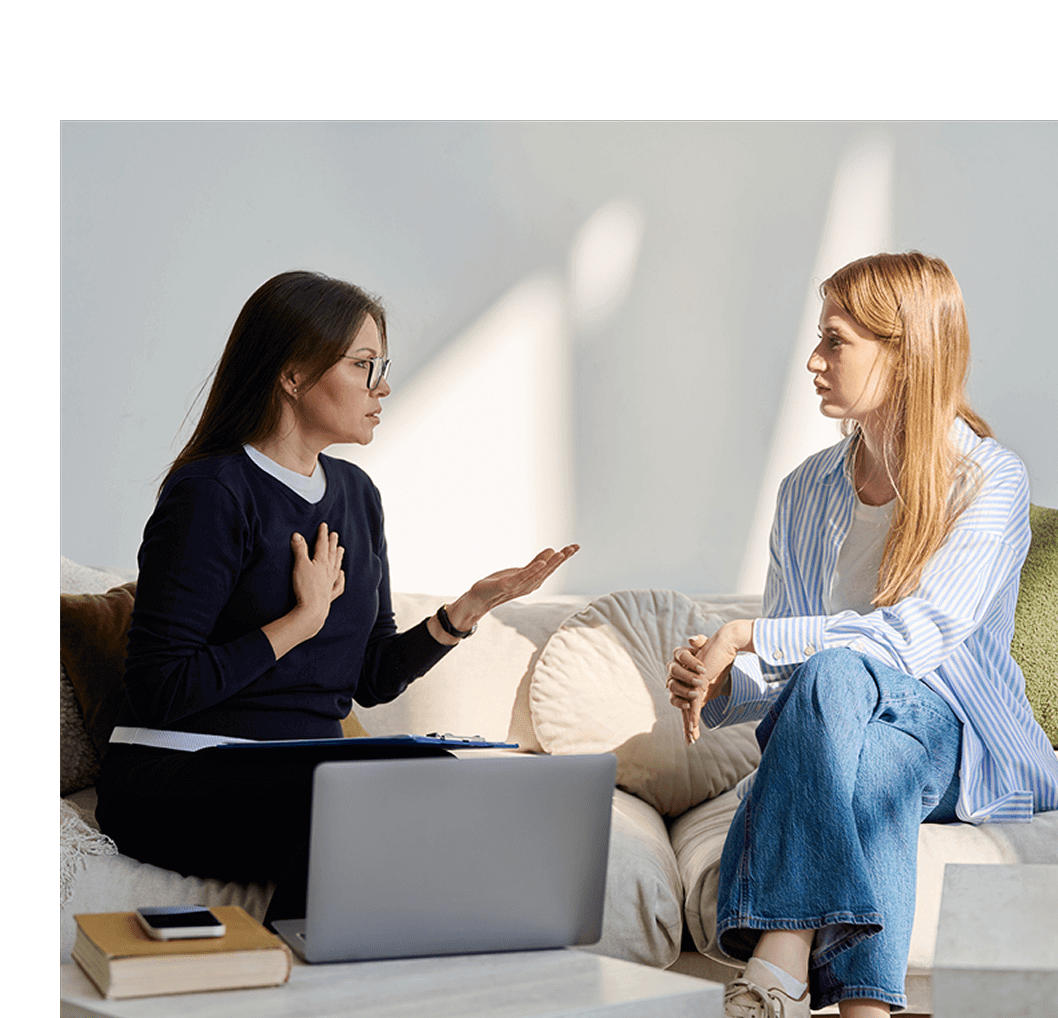 Two women engaged in a serious conversation indoors with a laptop nearby.